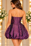 A woman shown from the back wearing a strapless short dress in shiny purple fabric with a structured corset-style back and a full gathered skirt. The background is softly blurred with warm lights and floral arrangements.