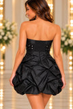 A woman shown from the back wearing a strapless black mini dress with a ruched embellished waistband and a voluminous gathered skirt, photographed in a softly lit indoor setting with blurred floral arrangements and warm lights.
